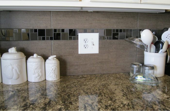 A kitchen counter with white decorative canisters and a utensil holder with various kitchen utensils. An electrical outlet and switch are visible on the tiled backsplash.